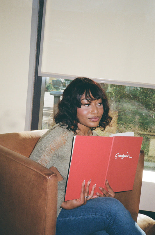 A woman in a chair, reading a book, wearing a human hair wig with stylish bangs.