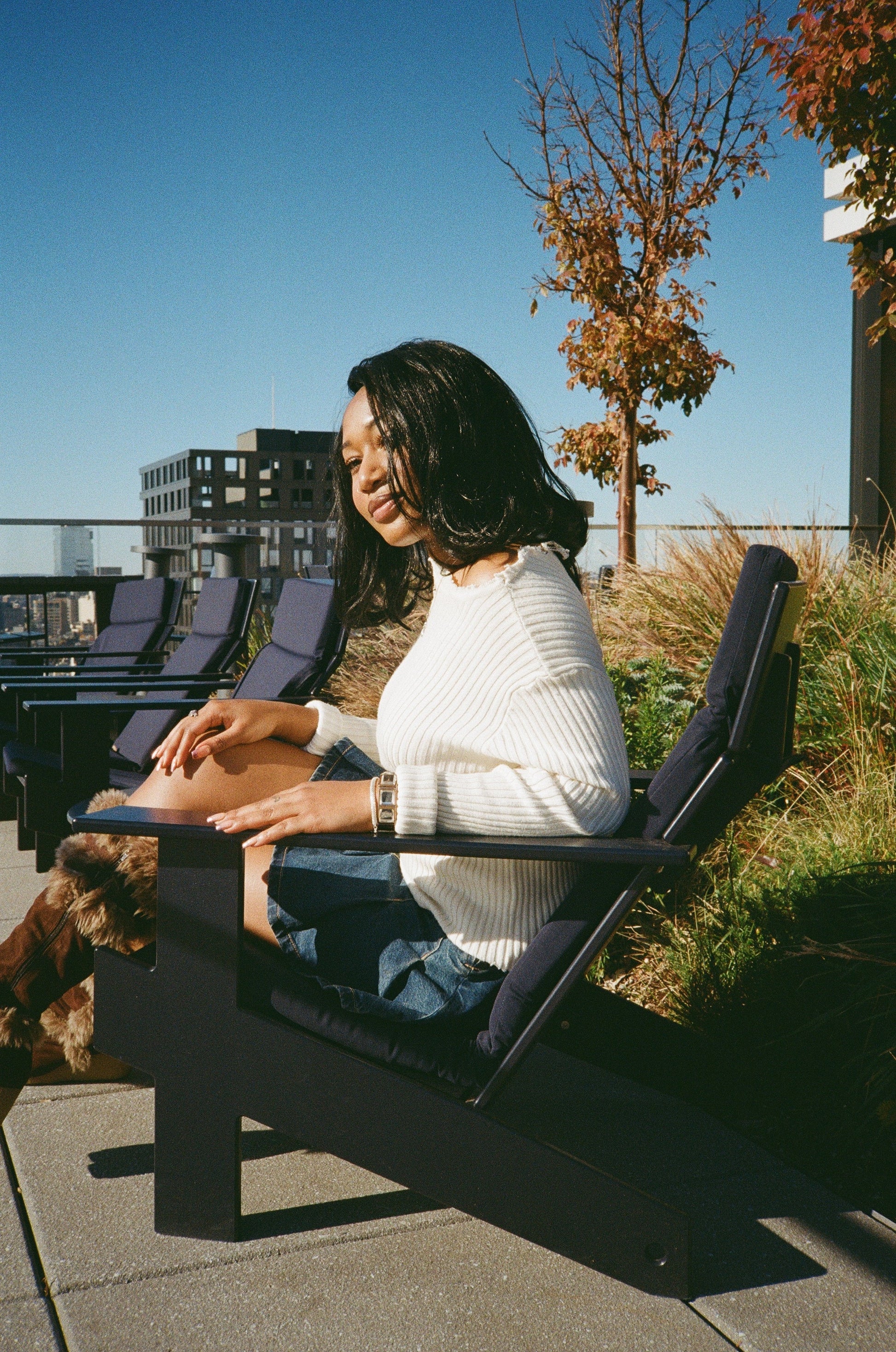 A stylish woman wearing a white sweater and blue skirt sits on a bench, her black synthetic hair wig with flipped ends completing her trendy outfit.