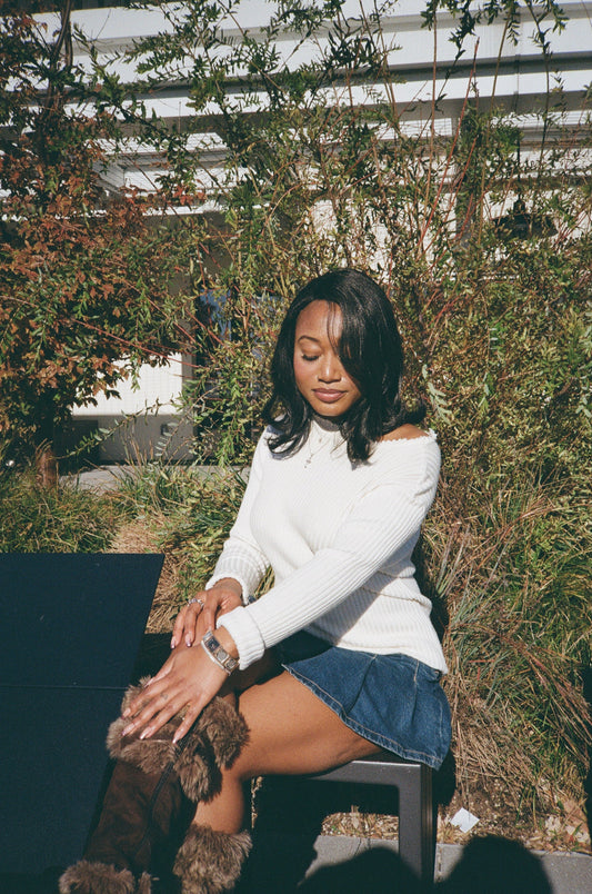 A stylish woman wearing a white sweater and blue skirt sits on a bench, her black synthetic hair wig with flipped ends completing her trendy outfit.